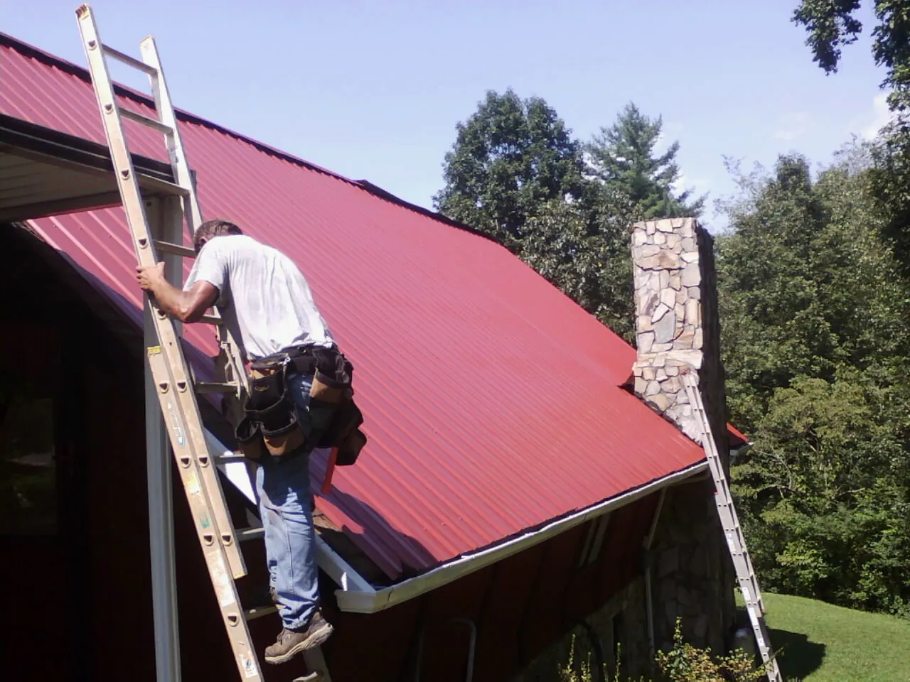 Roofer on a ladder beside a red metal roof and stone chimney — Soffit & Fascia Repair by Roofing Brothers in Palco