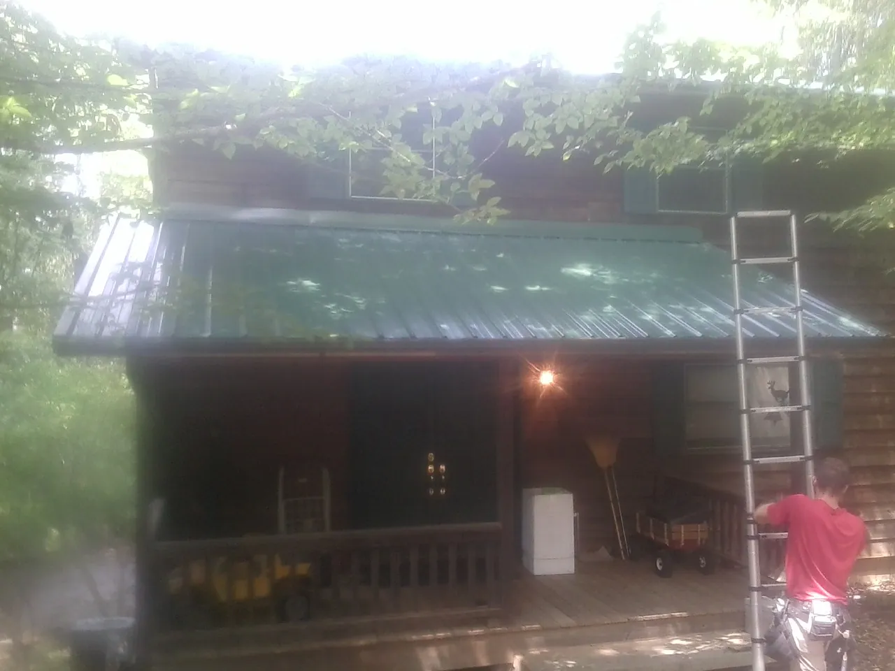 Worker on a ladder at the green porch roof of a wooded cabin in Palco